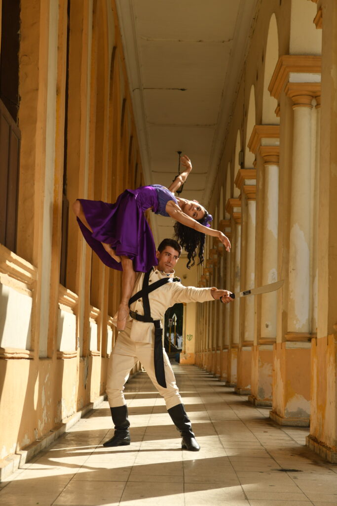 A male ballet dancer stands in a daylit hallway. He holds a sword out in front of him and in his right hand he holds up a female ballet dancer who is suspended in the air. He is supporting her by the small of her back and she is draped over, with her arms outstretched into the air. She is wearing a purple dress.