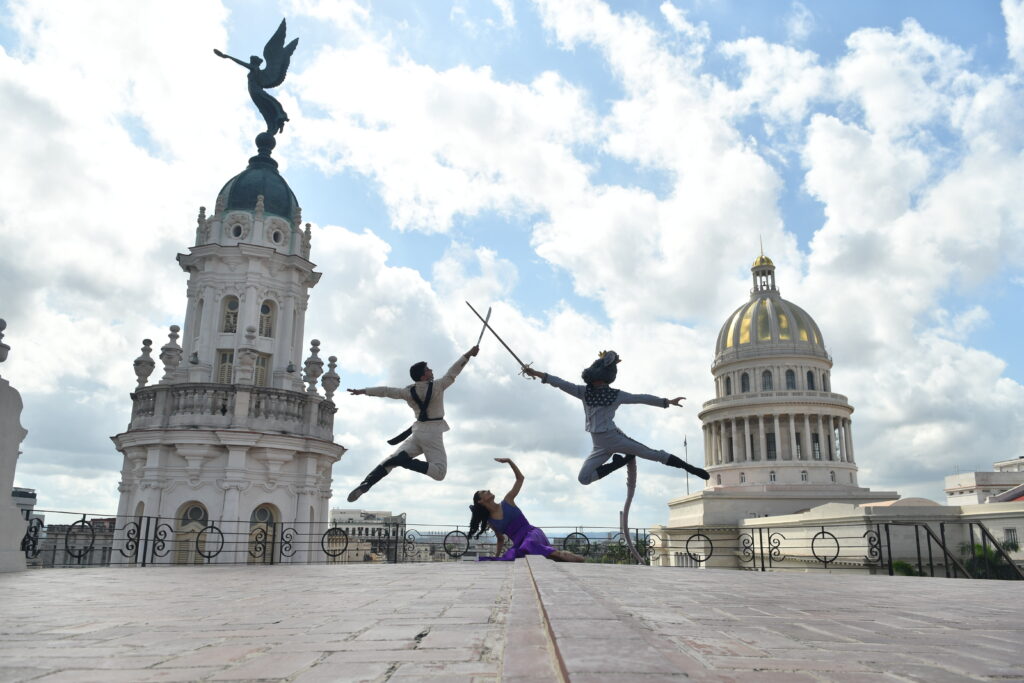 This image is on the rooftop of an old building overlooking Havana, the sky is blue. Two ballet dancers are mid-air , clashing swords in the middle. Each one has their free arm outstretched behind them and their legs elegantly outstretched too. One is a male soldier in a cream suit. The other is a rat king in a grey suit, but with a rat's tail and head, and a crown. A female ballet dancer sits on the floor in between them, cowering downwards with her hand held protectively above her and looking directly up. She is wearing a purple dress.