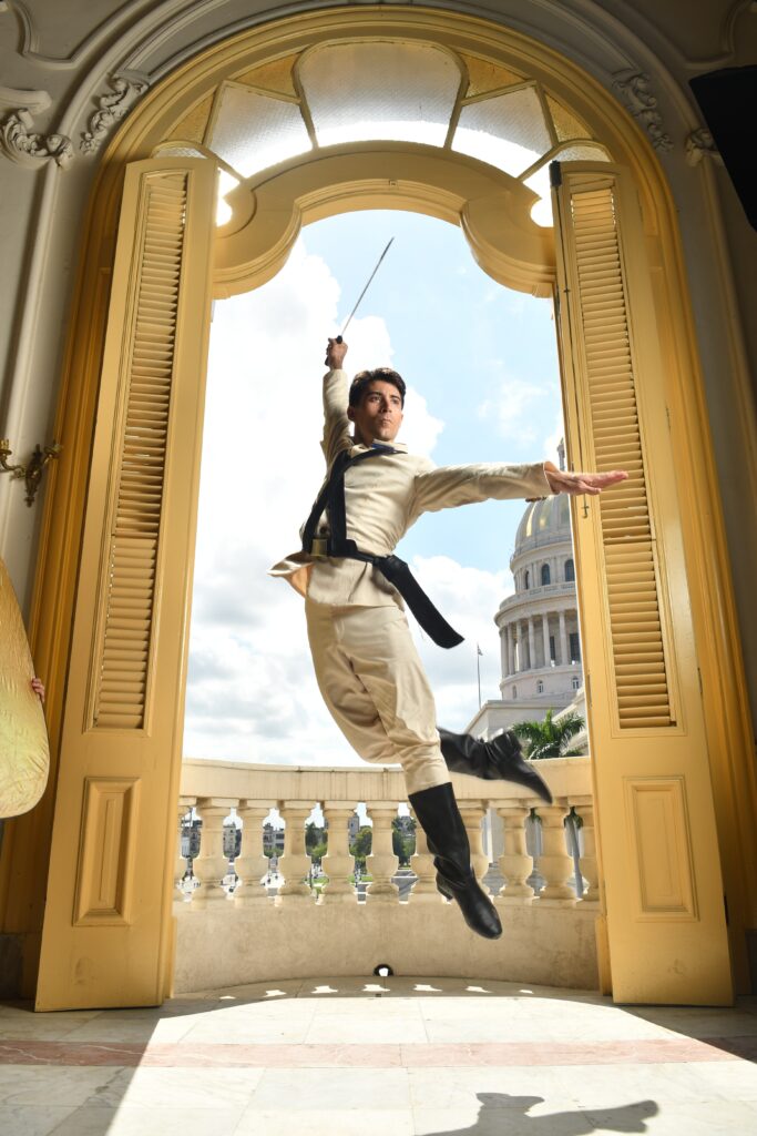 A male ballet dancer is jumping elegantly into the air in the doorway of a balcony. The double doors are open and looking out over a sunny Havana. The dancer has his left arm outstretched at a right angle to his torso, and his other arm is up in the air behind him and holding a sword.