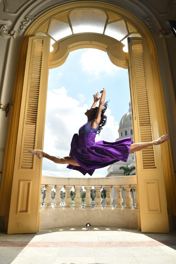 A female ballet dancer is jumping elegantly into the air in the doorway of a balcony. The double doors are open and looking out over a sunny Havana. The dancer is in side on and in the splits mid-air. Her arms are raised elegantly above her head and she is looking proudly upwards. She is wearing a vivid purple dress and ballet shoes.